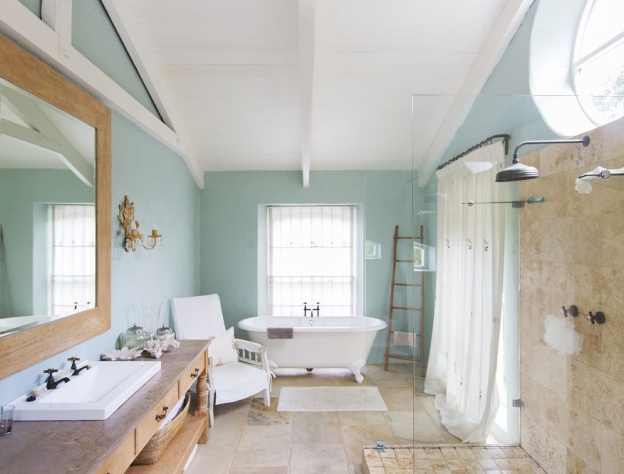 Lifestyle image of a farmhouse style bathroom, with wooden floors and turquoise walls, a walk in shower enclosure, wooden washstand, white freestanding bath with painted legs and white curtains across the windows